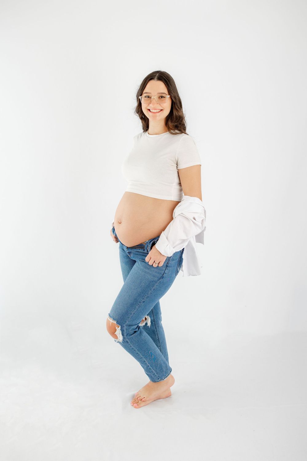 Woman in casual white top and blue jeans poses against white background in minimalist studio photoshoot.