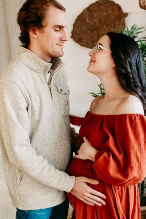 A couple in a romantic embrace wearing a beige sweater and red dress against a neutral background.