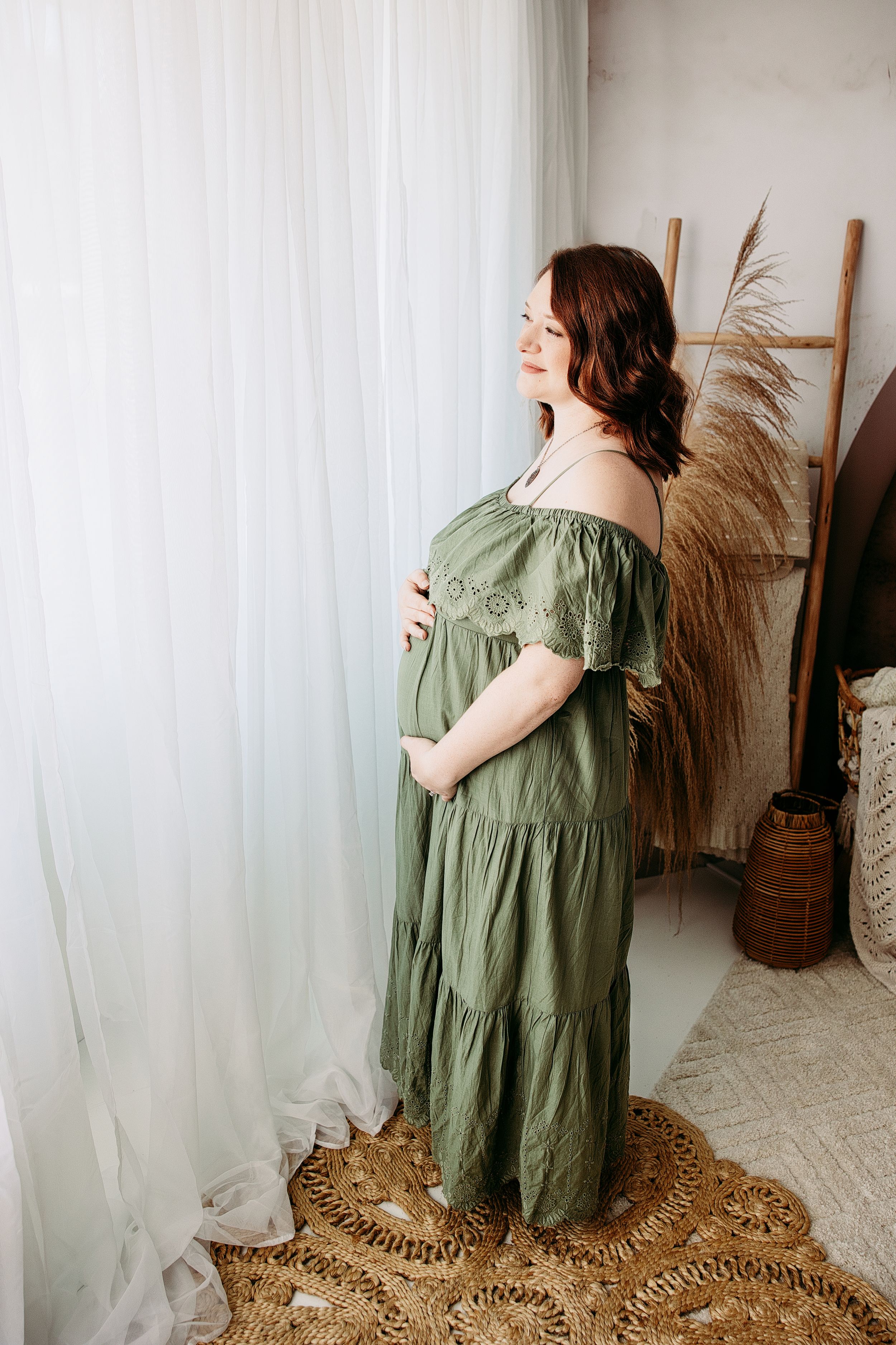 Woman in flowing olive green dress poses in front of white curtain and natural decor. Middle TN maternity photographer