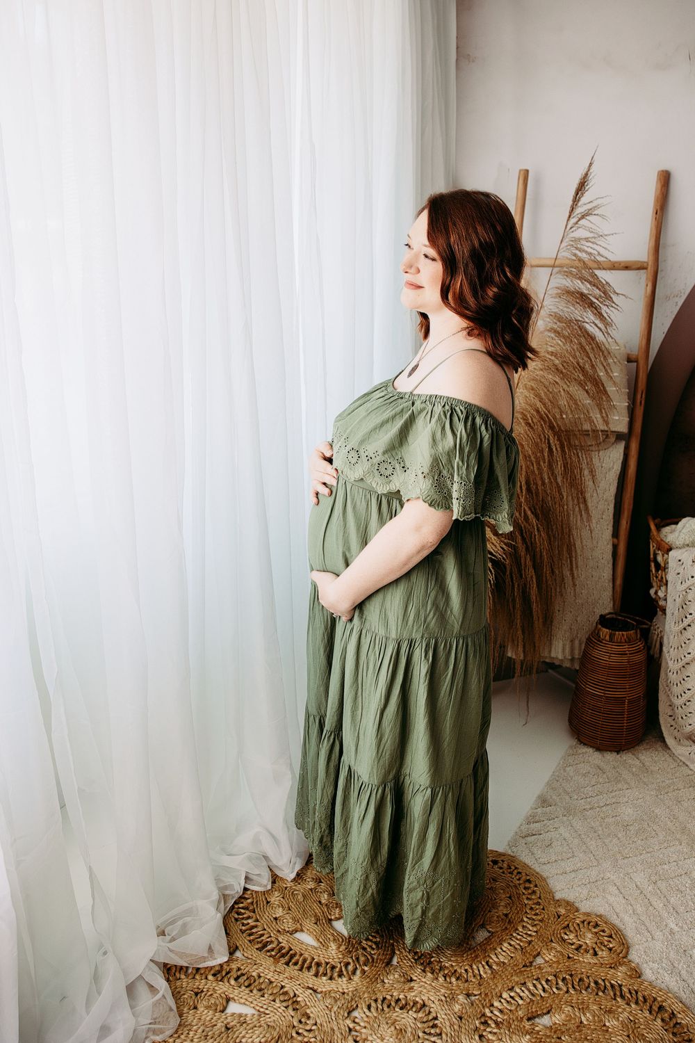 Woman in flowing olive green dress poses in front of white curtain and natural decor. Middle TN maternity photographer