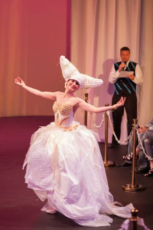 Performer in elaborate white gown and tall headpiece dances gracefully on stage.
