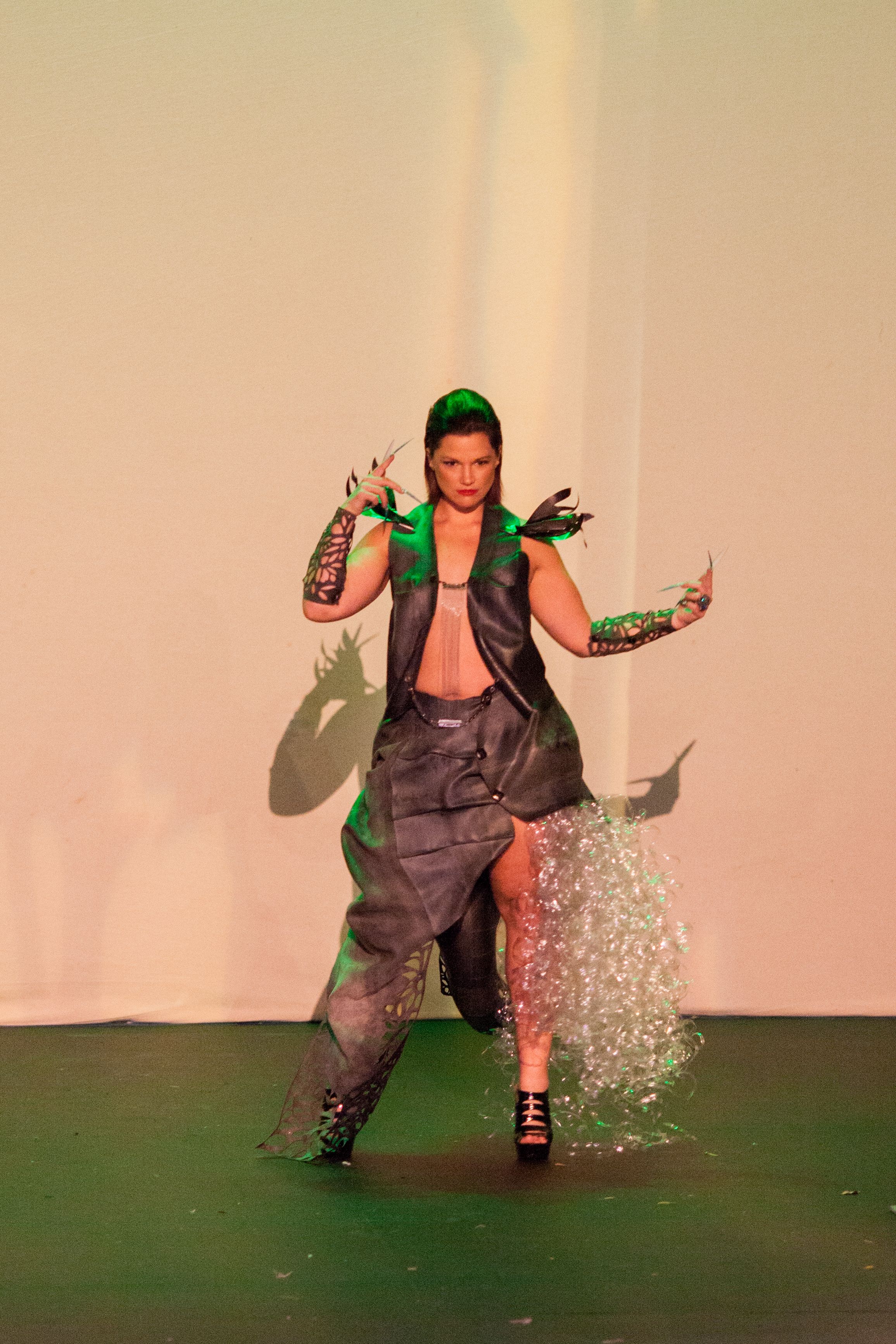 Performer in edgy costume with green lighting performs dynamic dance move on stage.