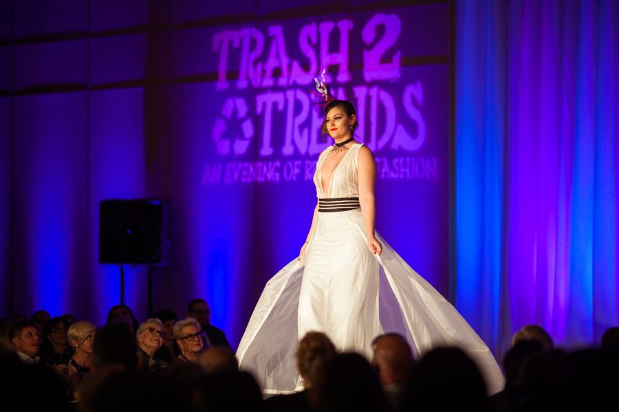 Model walks runway in elegant white ballgown at Trash 2 Trends recycled fashion show with purple lighting.