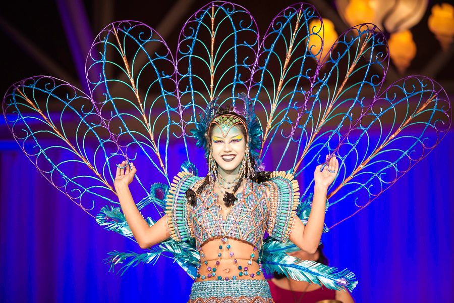 Performer wearing elaborate peacock-inspired costume with blue LED feathers on illuminated stage.