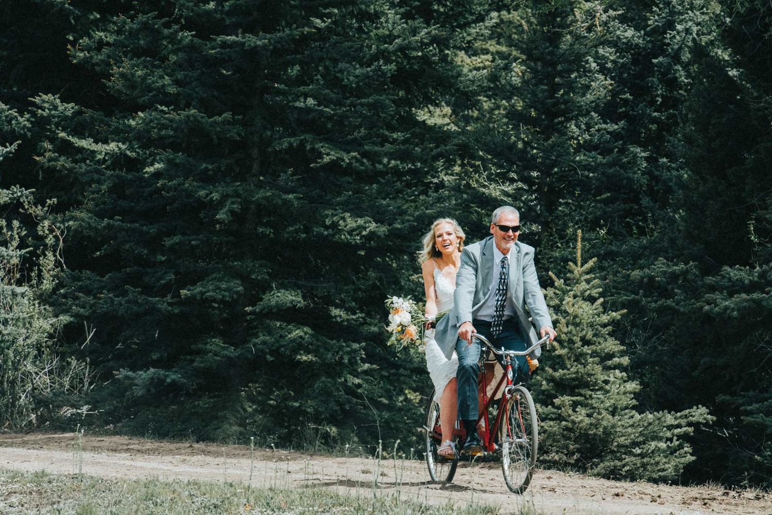 A couple rides a bicycle together through a wooded path on a sunny day.
