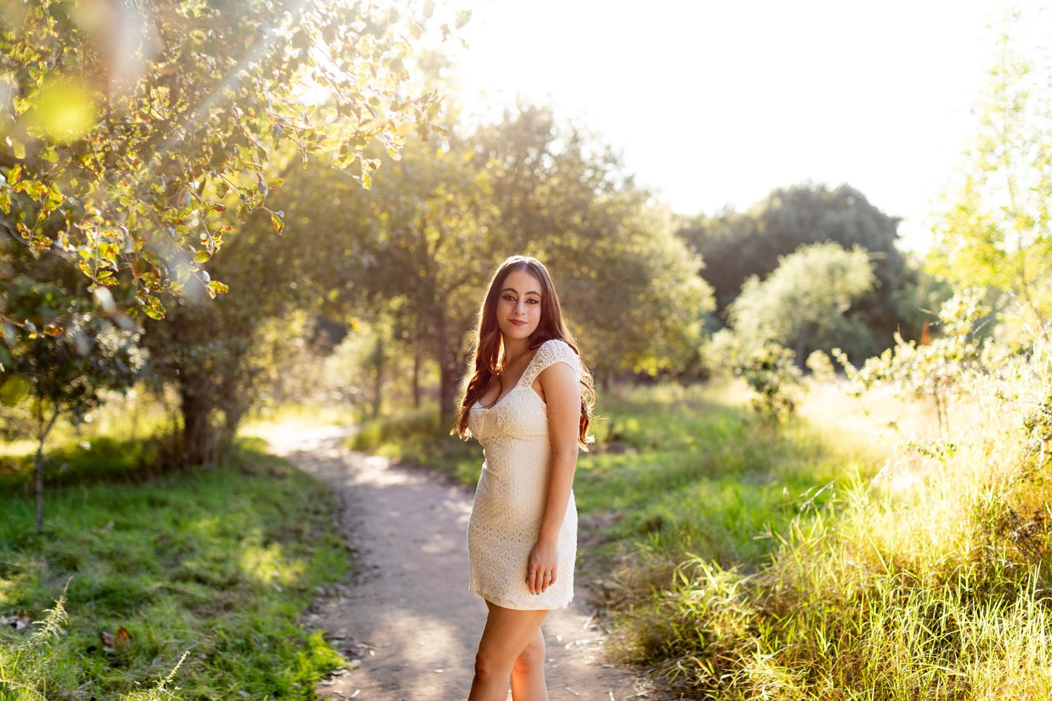 A high school senior in a white dress stands on a sunlit path through a lush garden setting during golden hour.