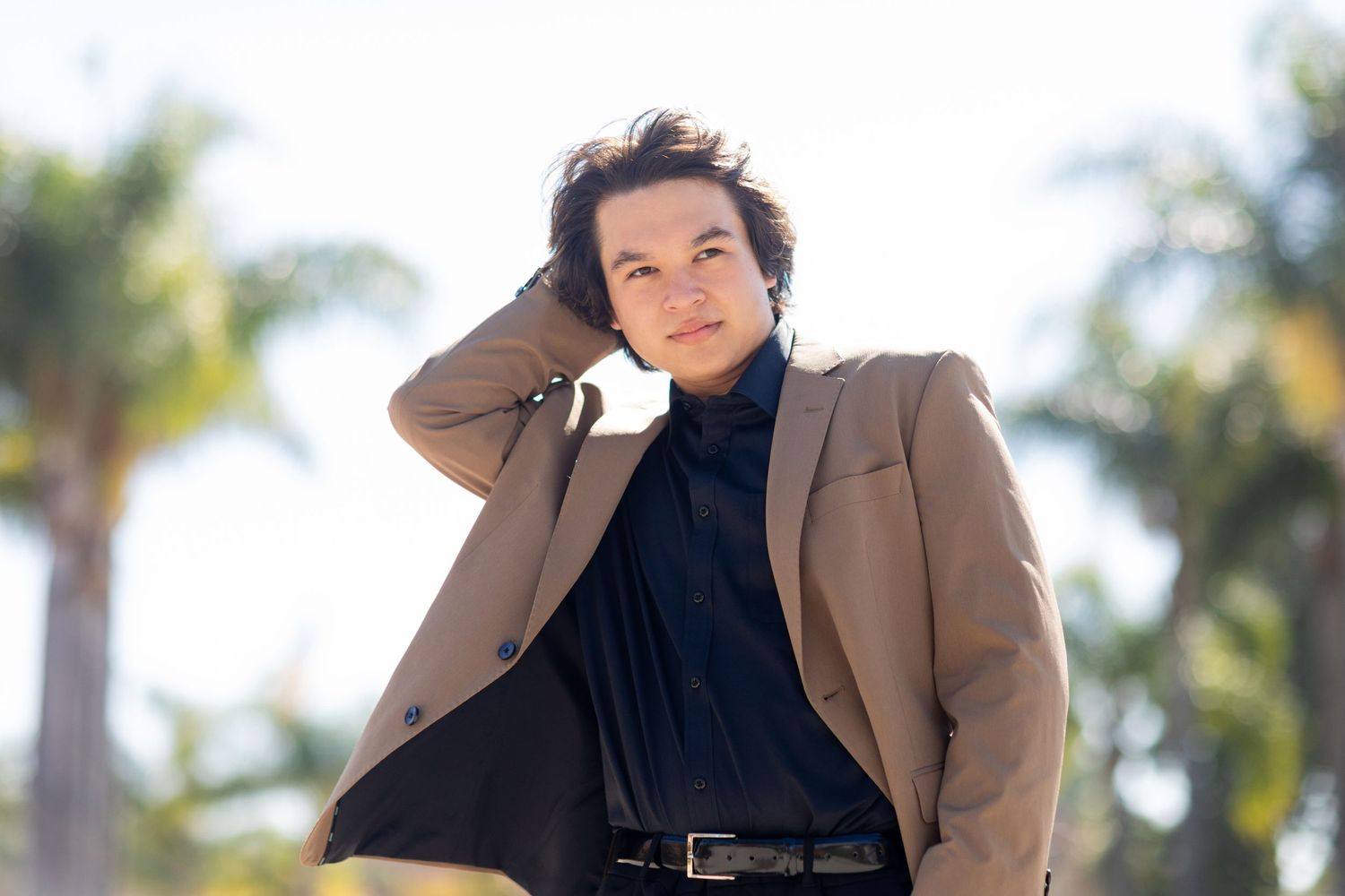 A high school senior in a tan blazer and black shirt poses outdoors against a palm tree backdrop on a sunny day.