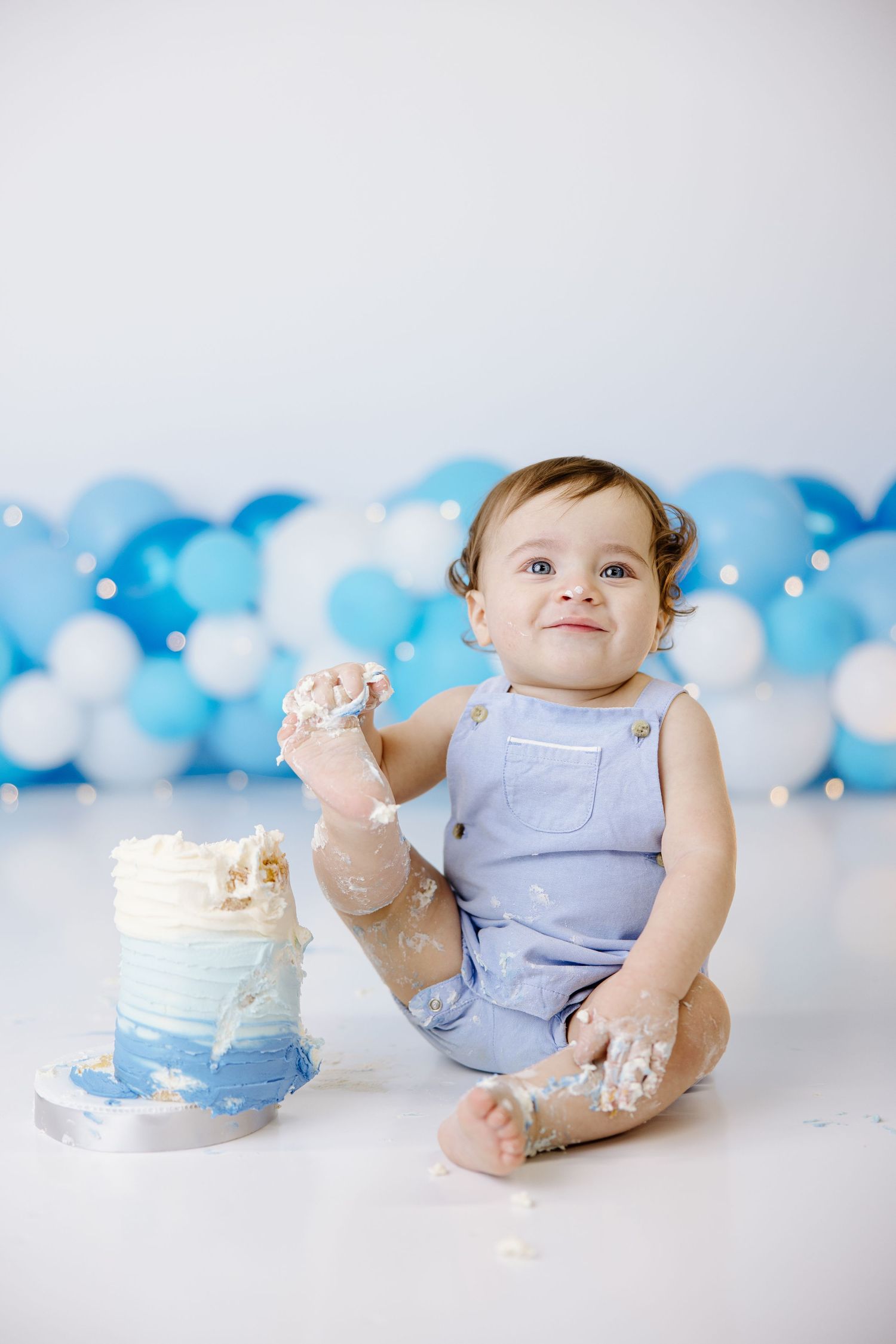 Baby in blue romper with cake smash photo setup against blue balloon backdrop.