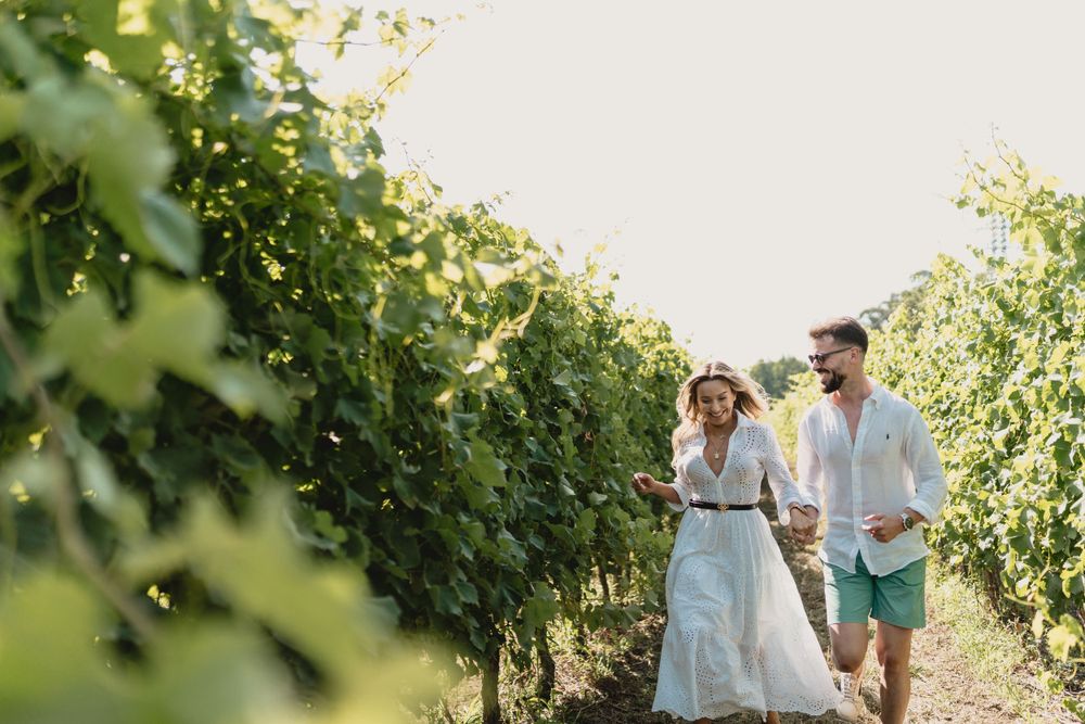 Couple walking through rows of vineyard on sunny afternoon holding drinks.