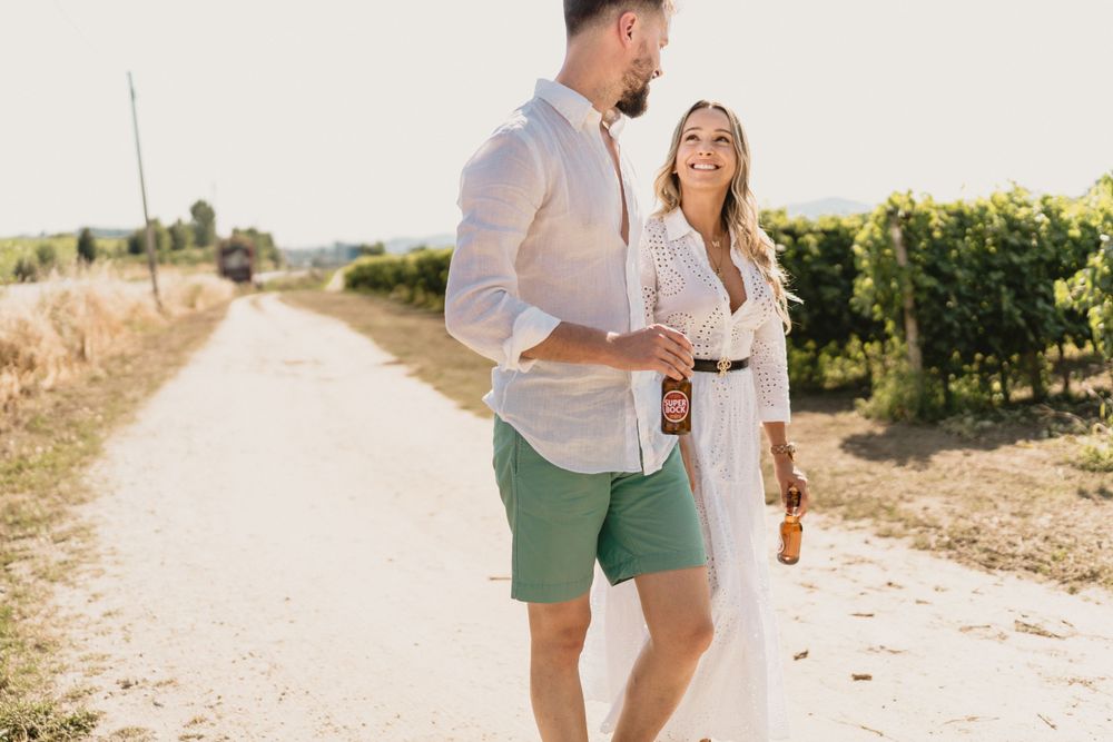 Couple walking together on sunny path through vineyard holding beverages.