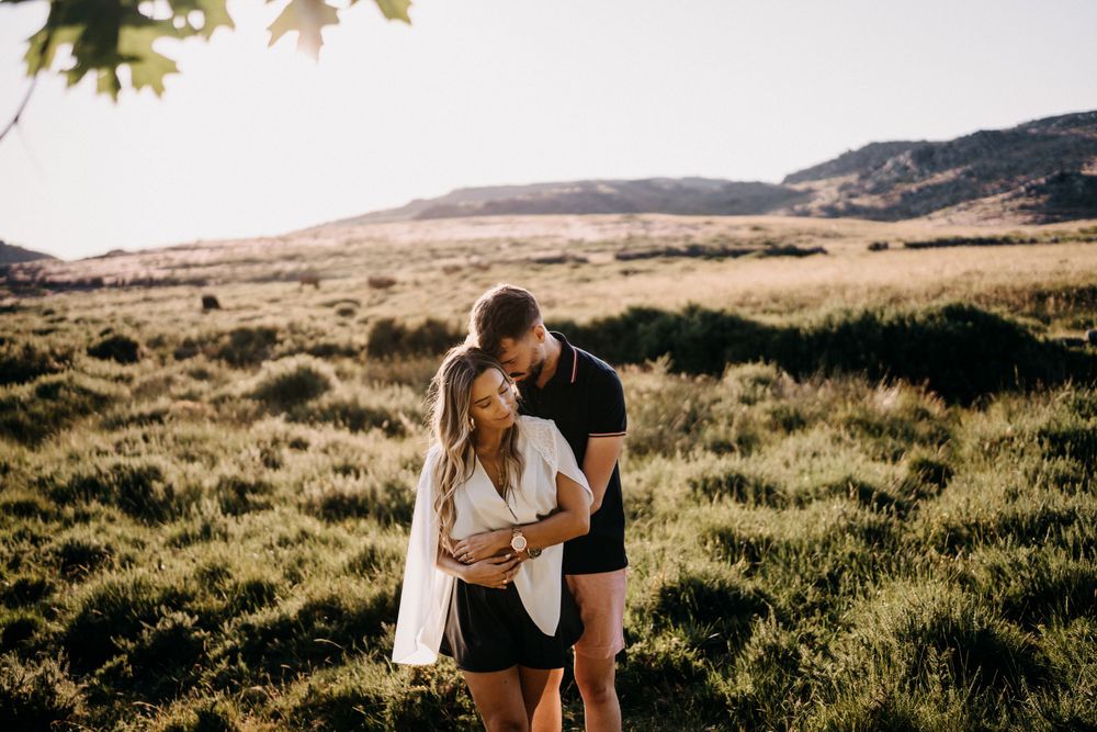 Couple embracing in a sunlit meadow with mountains in the background.