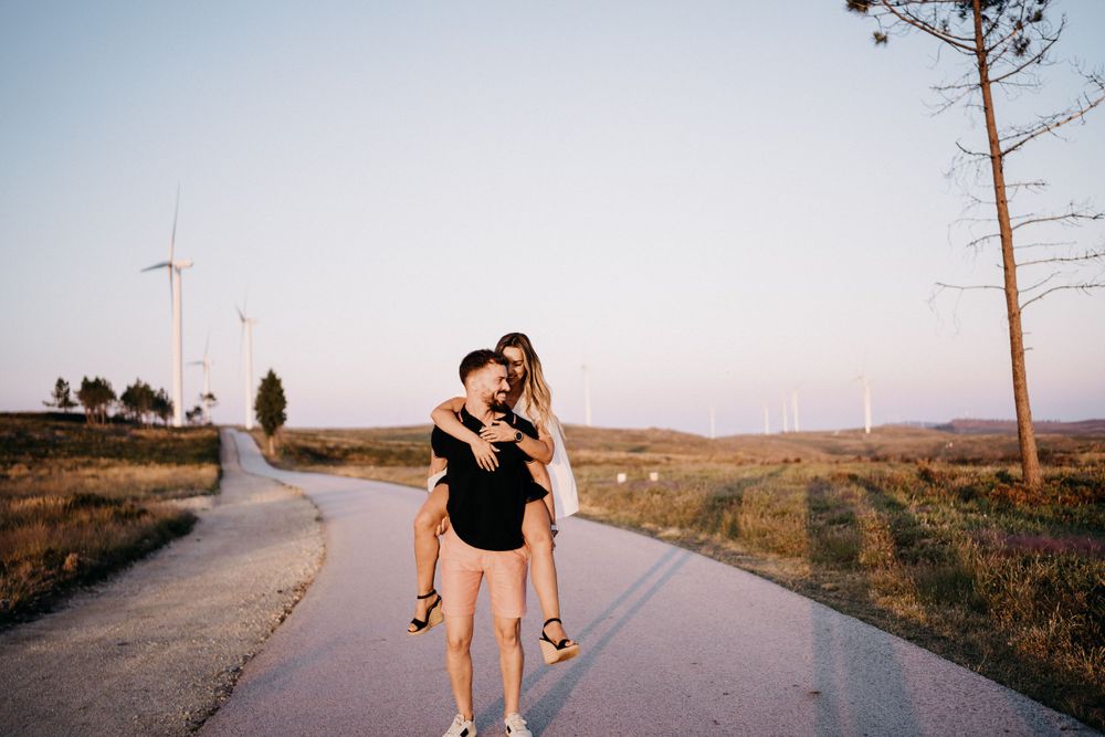 Playful piggyback pose on empty road with wind turbines in distance.