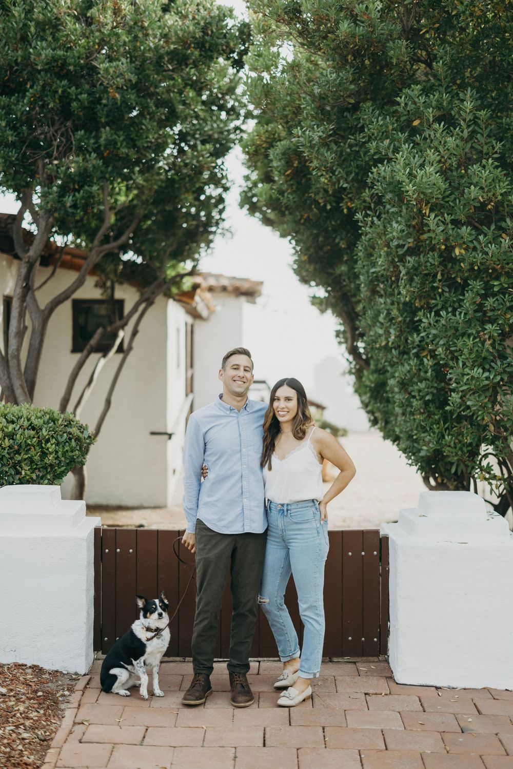 A couple poses with their black and white dog against a white stucco wall and brown wooden gate.