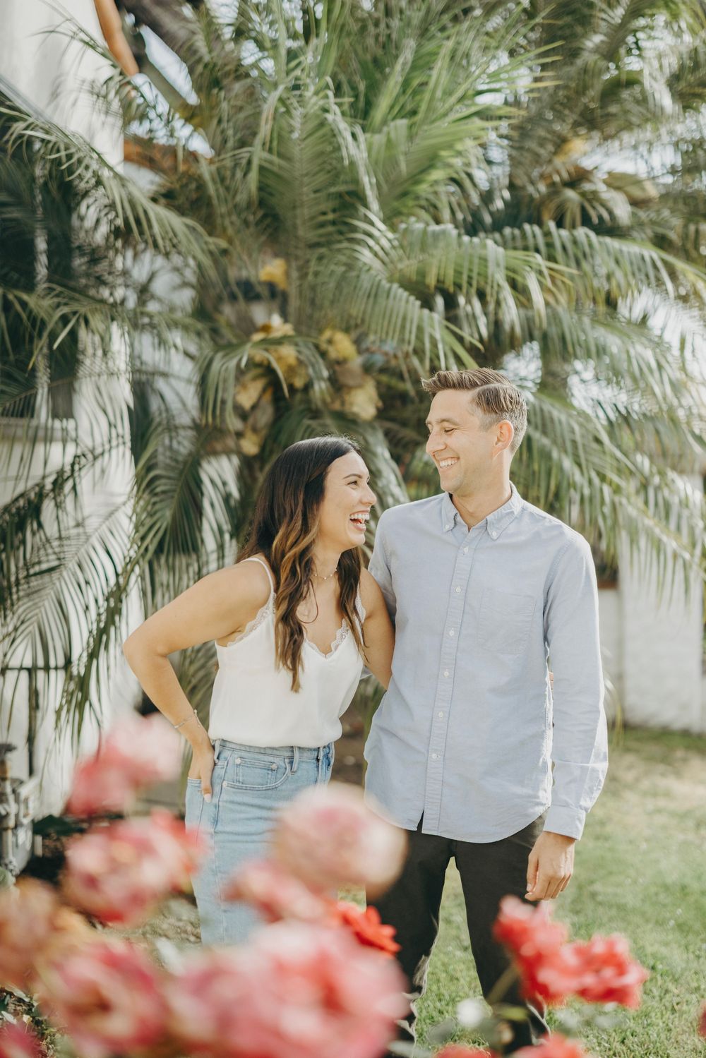 A couple shares joyful laughter among tropical palm trees and pink flowers in a garden setting.