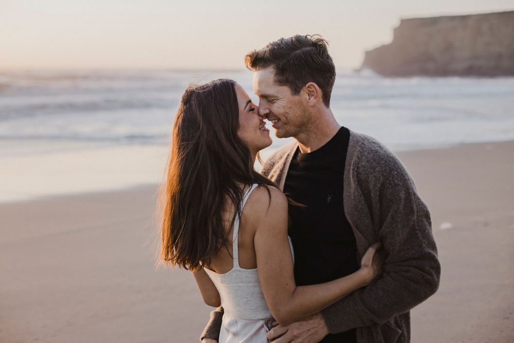 Two people embrace on a beach at sunset with coastal cliffs in the background.