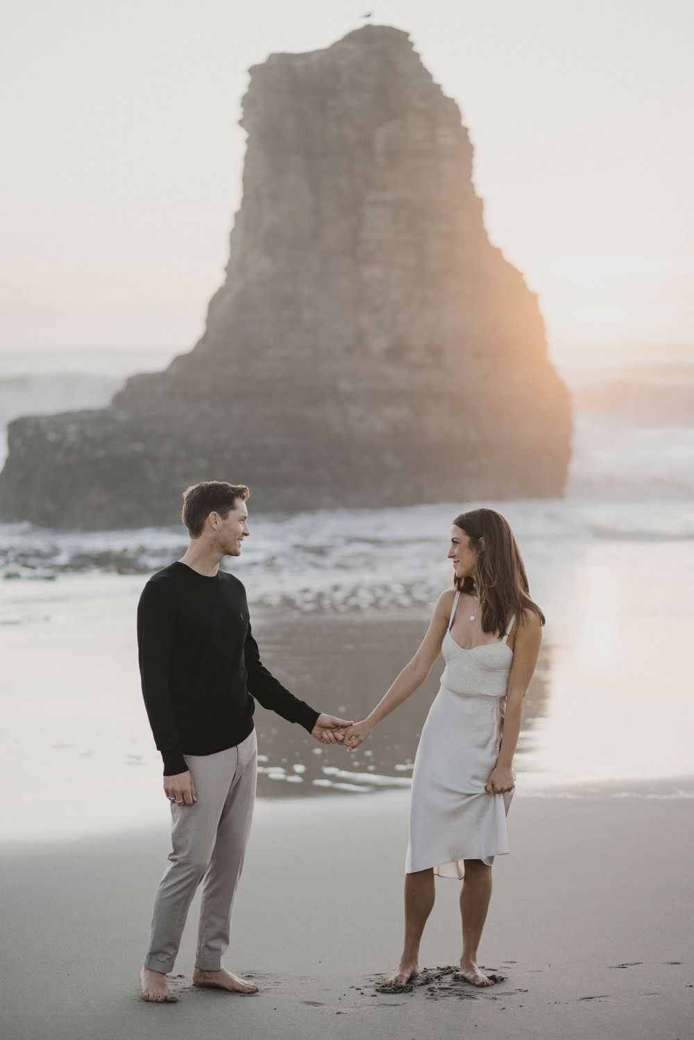 A proposal scene unfolds on a sandy beach with dramatic rock formations and sunset in the background.