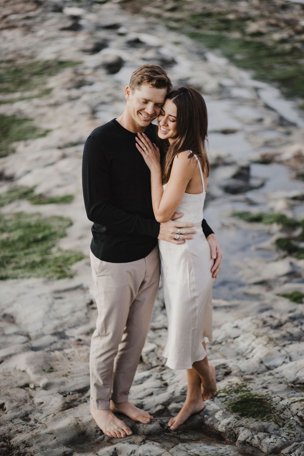 A couple shares an intimate moment on rocky coastal terrain with ocean views.