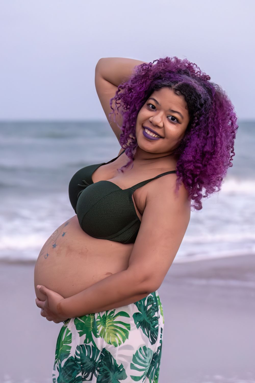 A joyful beachside maternity portrait with vibrant hair and tropical outfit.