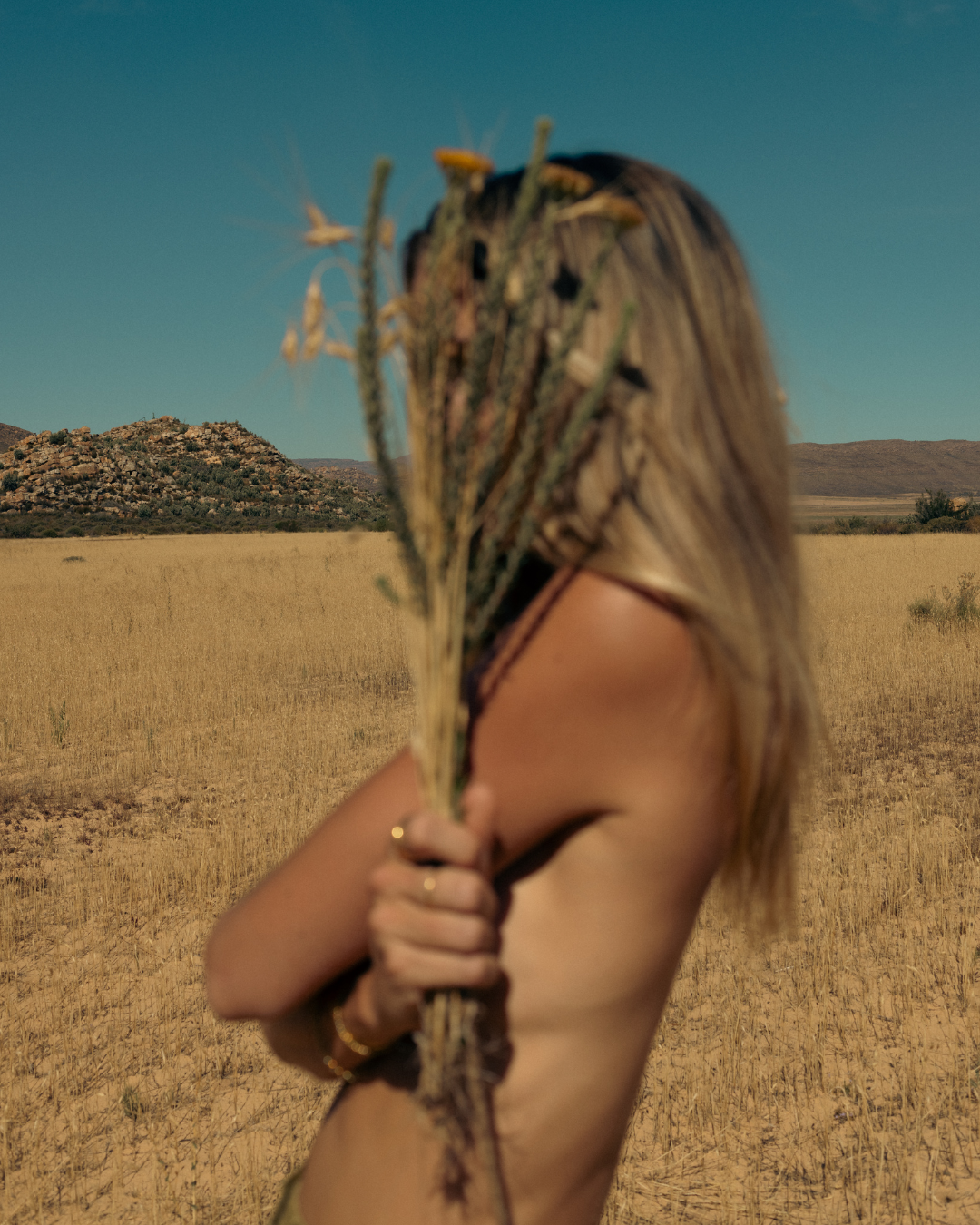 Person holding dried flowers against their face in a golden wheat field at sunset.