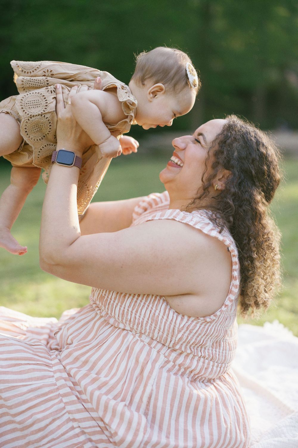 Joyful outdoor portrait of parent lifting baby wearing beige eyelet dress in afternoon sunlight.