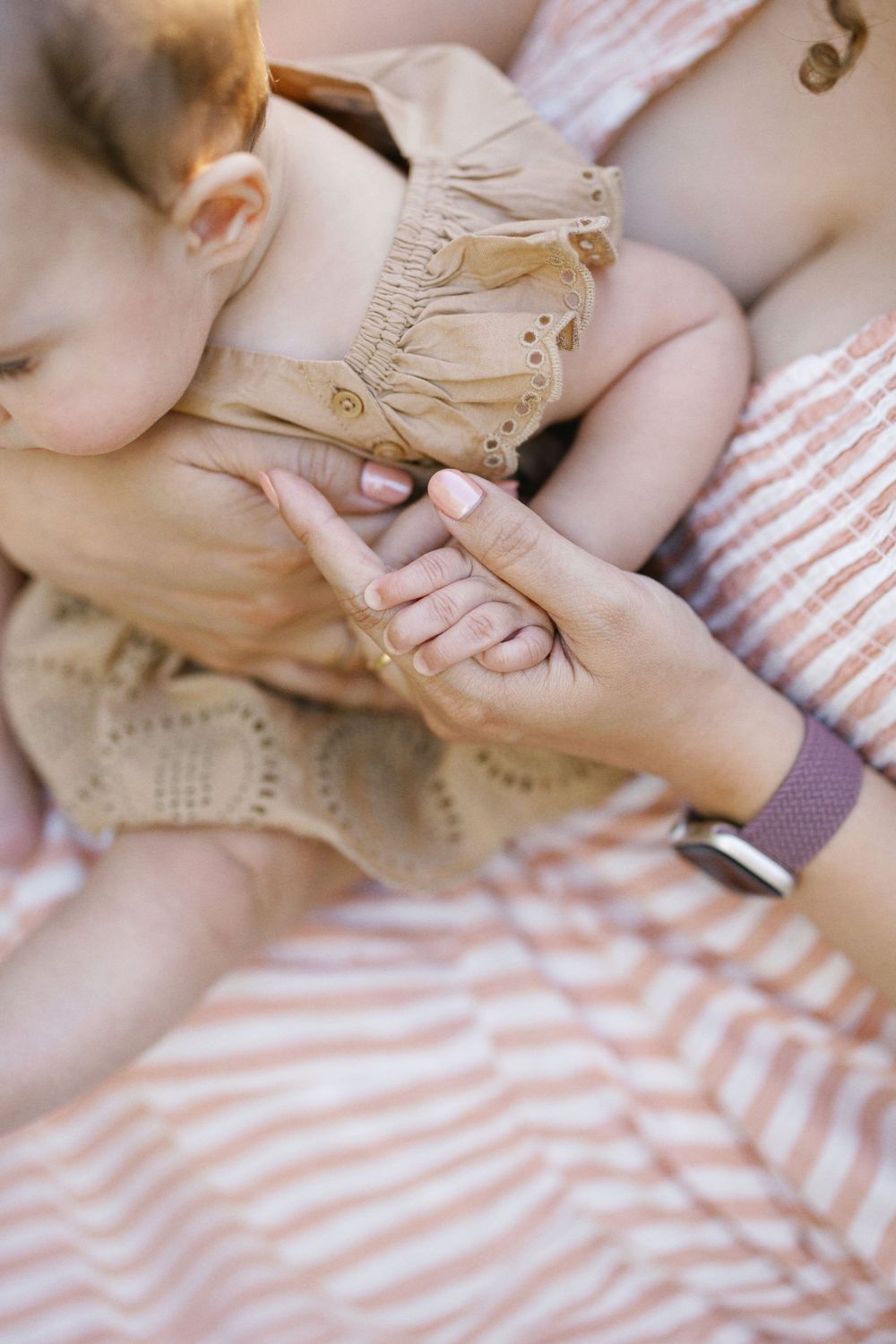 Close-up detail of hands holding baby wearing tan eyelet dress against striped fabric.