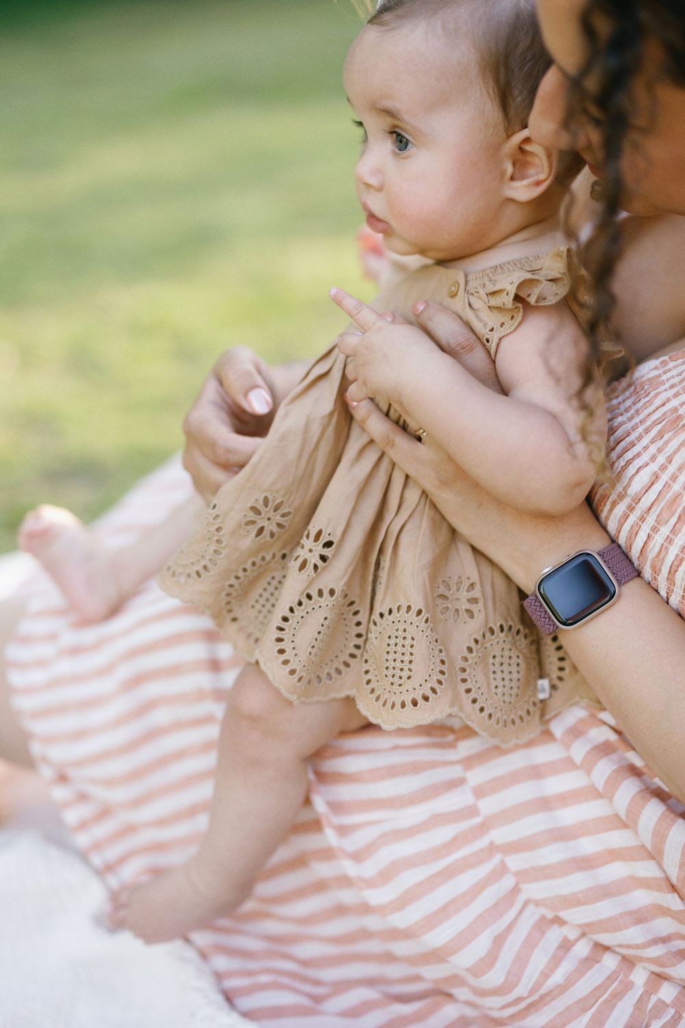Profile view of baby in tan eyelet dress seated with parent outdoors.