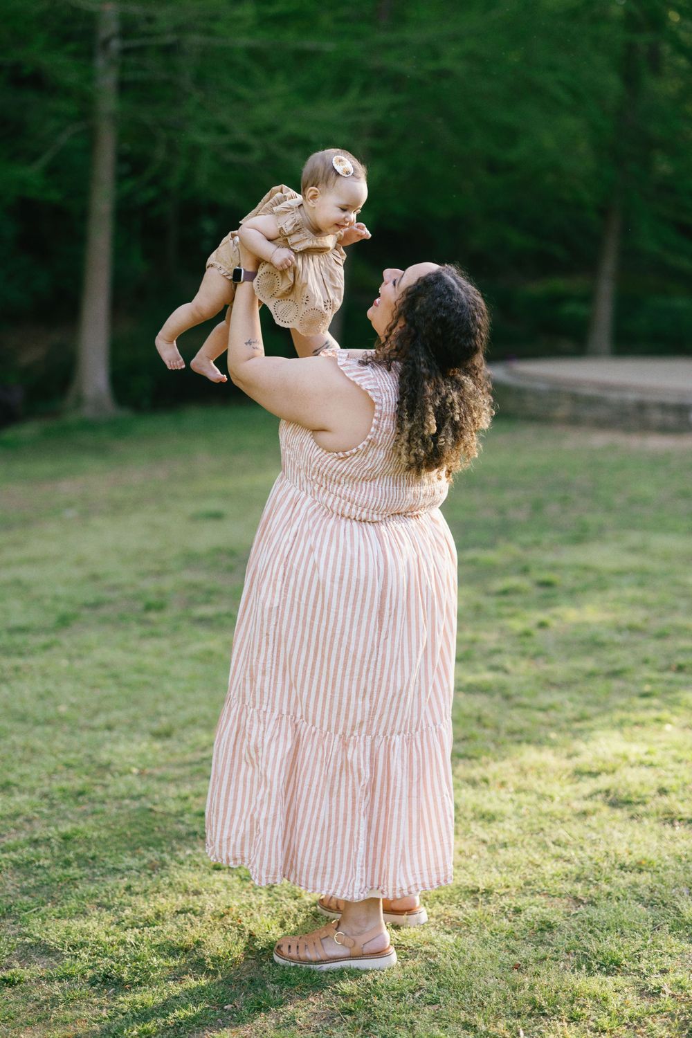 A tender moment outdoors as a parent lifts a baby wearing a beige dress against a green forest backdrop.