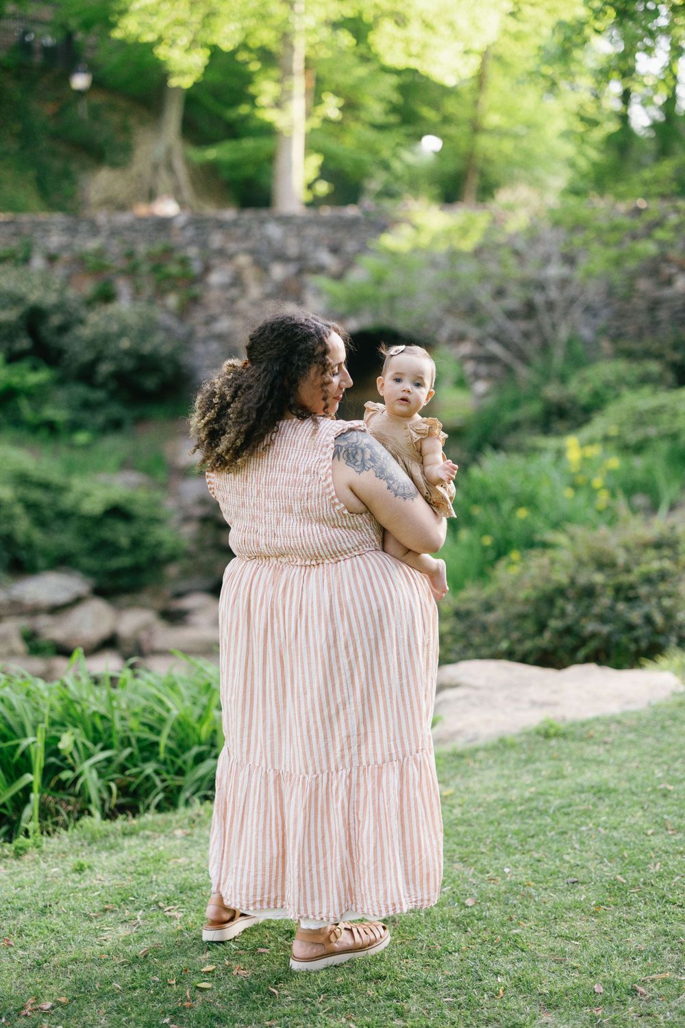 Parent and baby share a joyful moment in a garden setting near a stone wall.