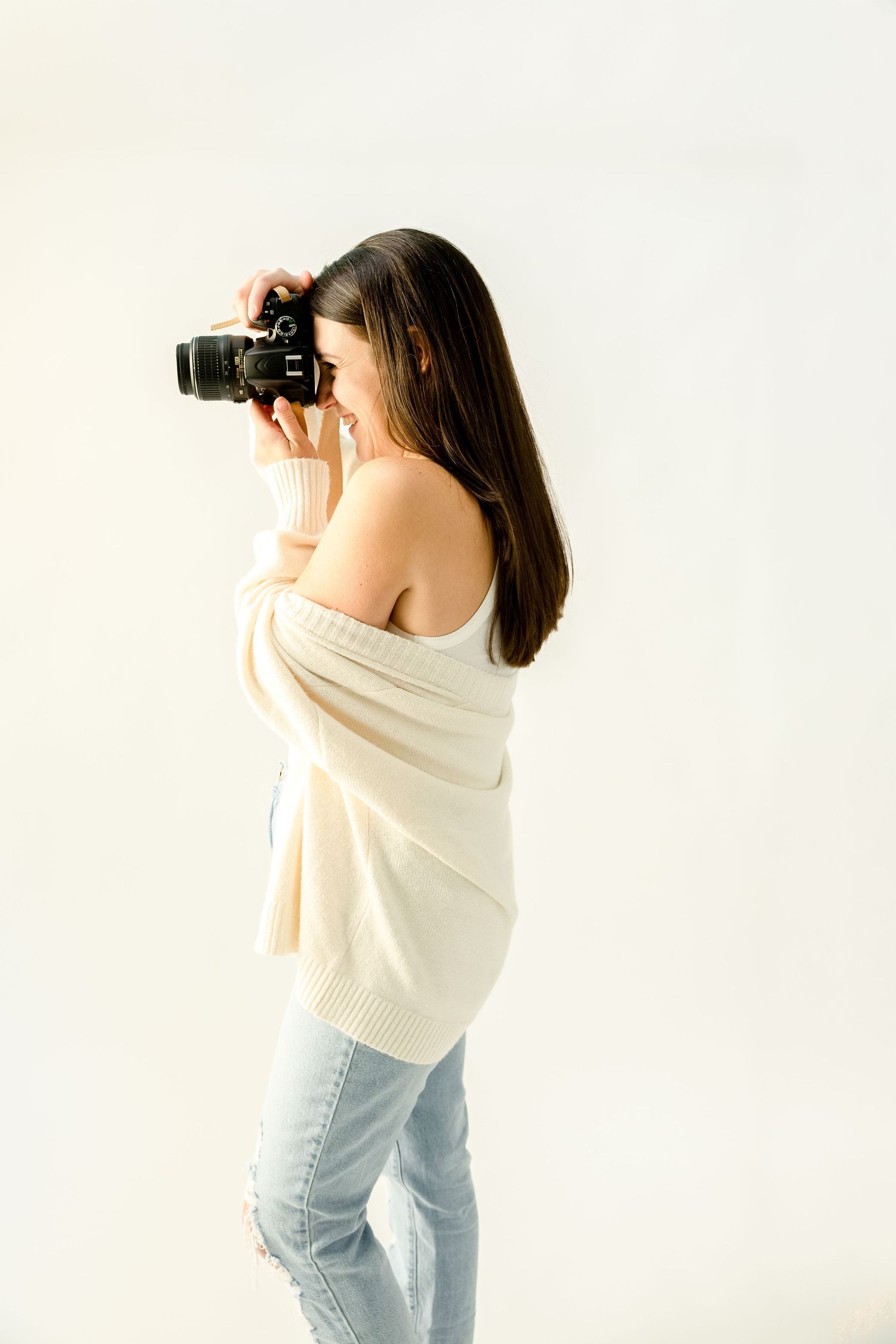 Photographer in cream sweater and jeans holding camera against white backdrop.