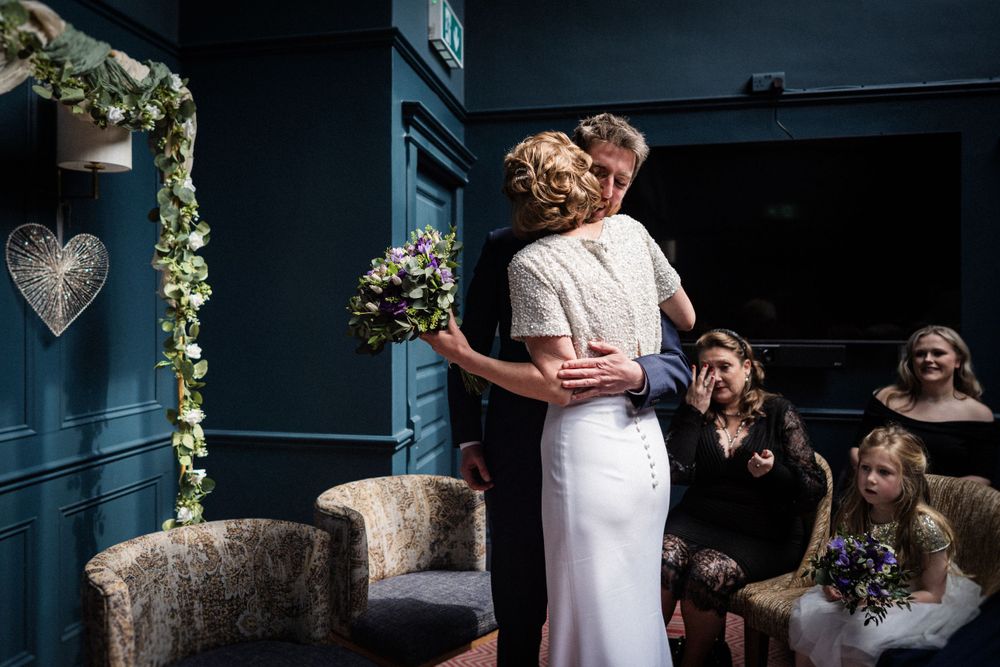 An intimate wedding ceremony moment captured in a room with navy blue walls and decorative floral garlands.