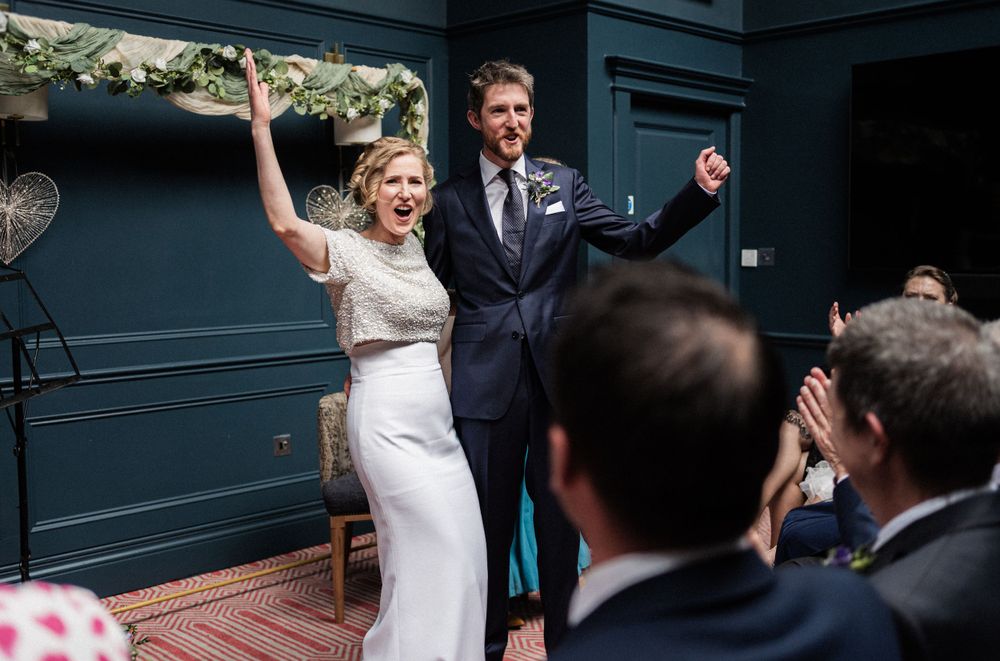 Newlyweds celebrate with raised arms during their wedding ceremony in a navy blue room with floral decorations.