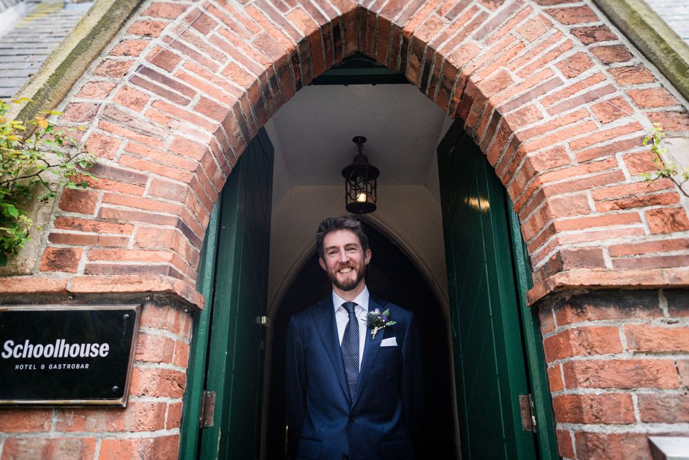 Groom standing in formal attire under a brick archway entrance of a schoolhouse.