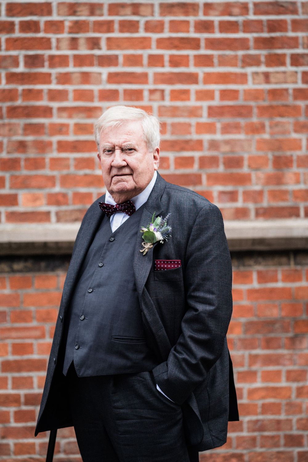 An elegant gentleman in formal attire with bow tie poses against a brick wall wearing a boutonniere.