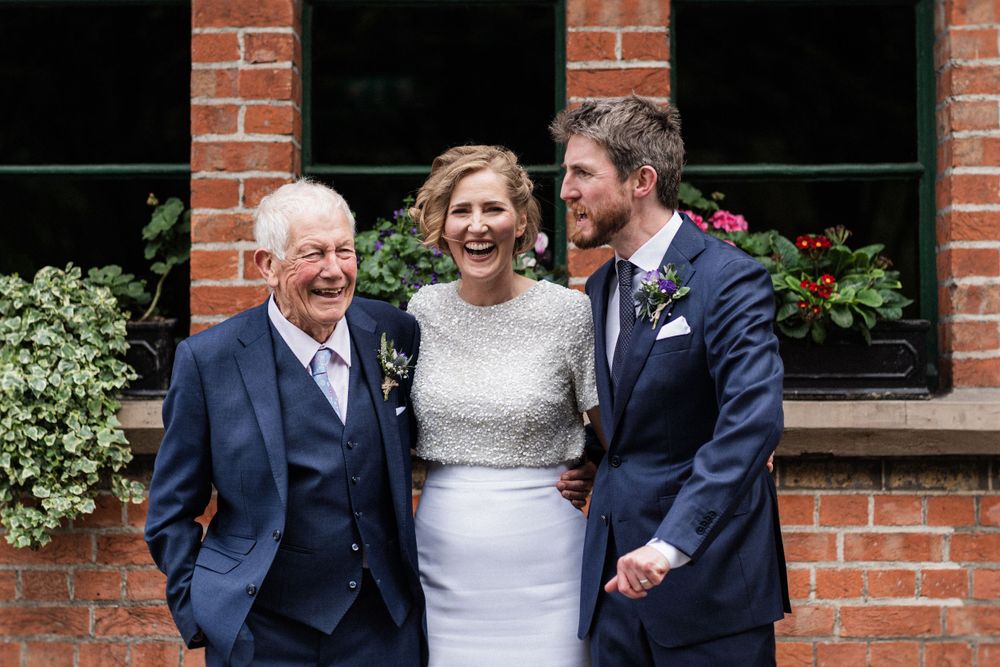 Three people share a happy moment together against a red brick backdrop at a wedding.
