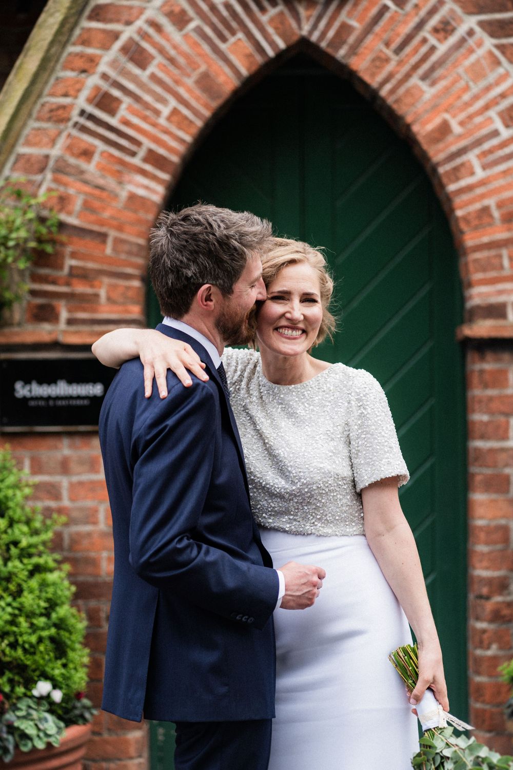 A couple shares an intimate moment in front of a green arched doorway with red brick.