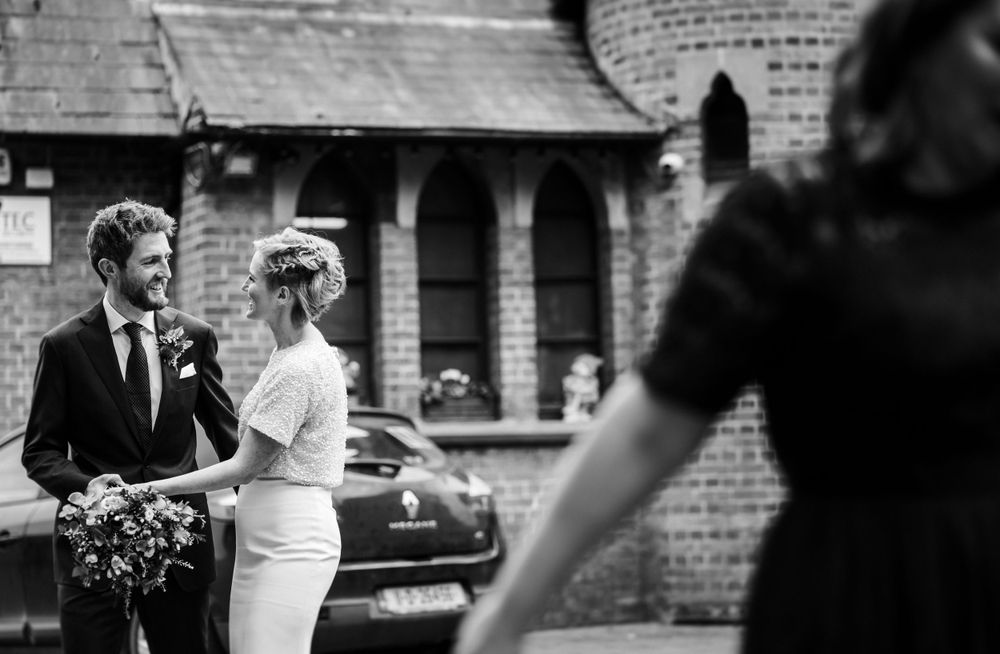 A black and white wedding scene with couple and onlooker against brick architecture.