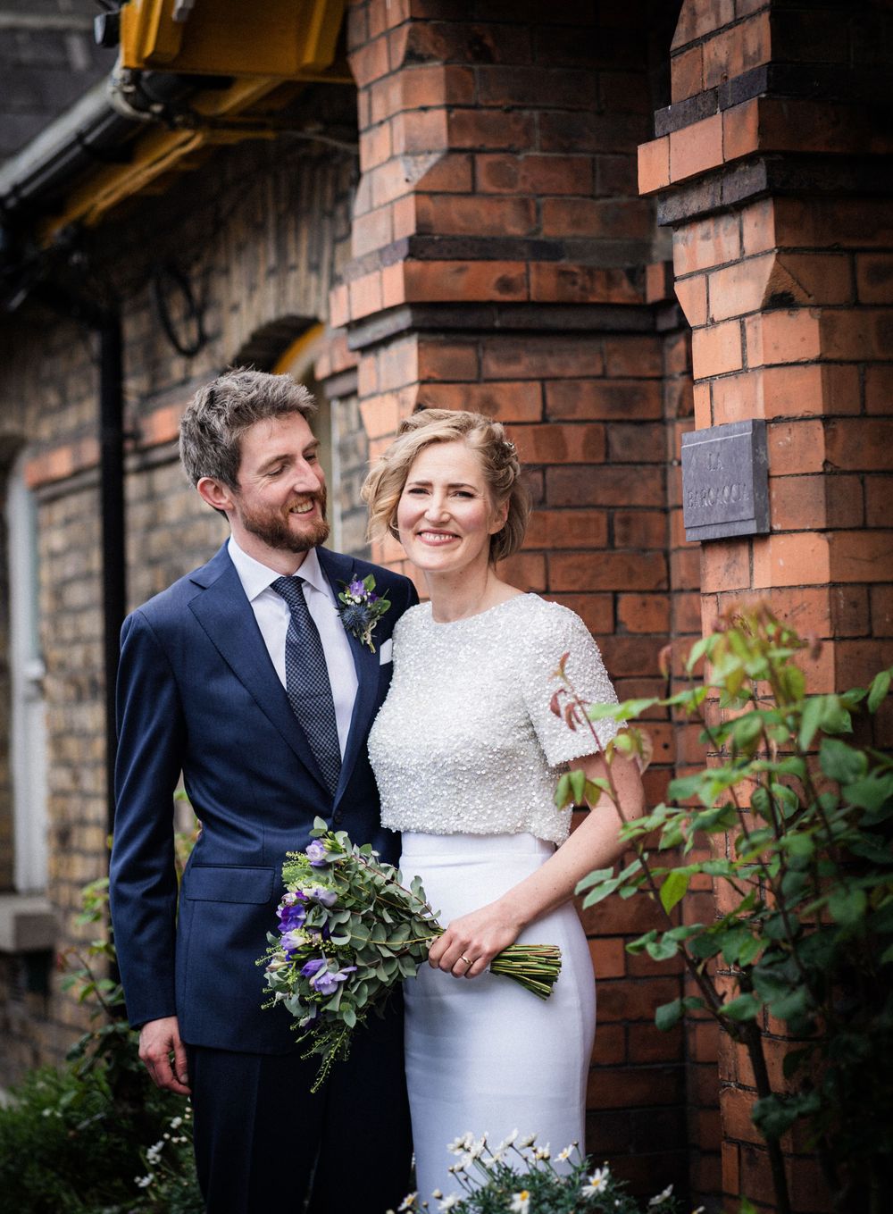 A couple in wedding attire smiles together against rustic brick pillars and greenery.