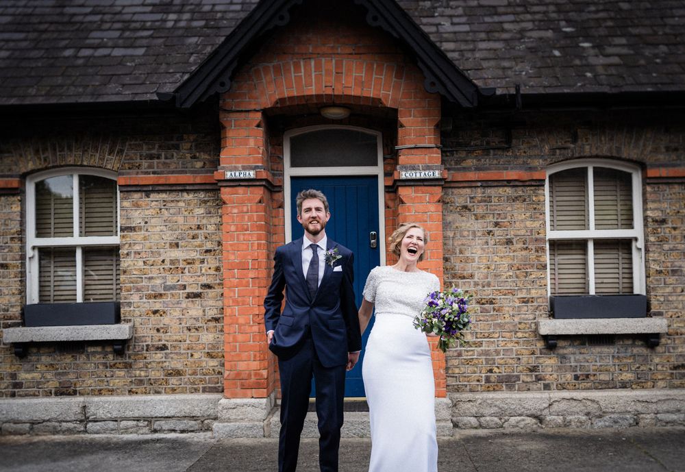 A couple stands in front of a historic brick building with a blue door on their wedding day.