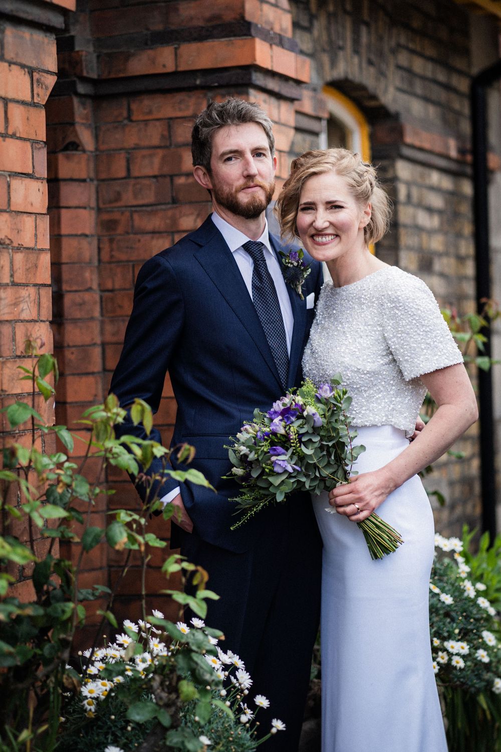 A newlywed couple poses in front of a rustic brick wall with purple floral bouquet.