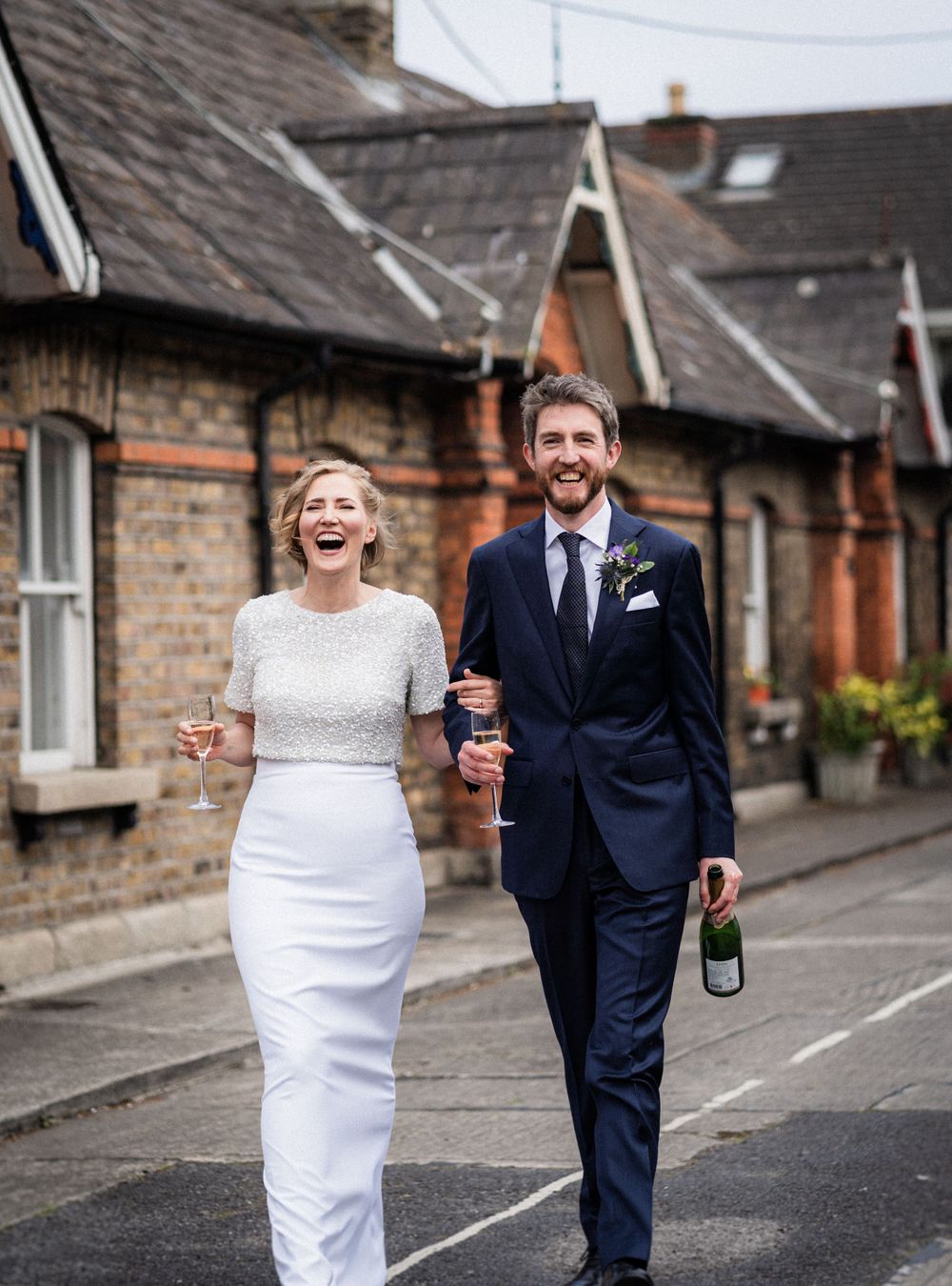 A joyful couple holding champagne bottles walks down a quaint residential street.
