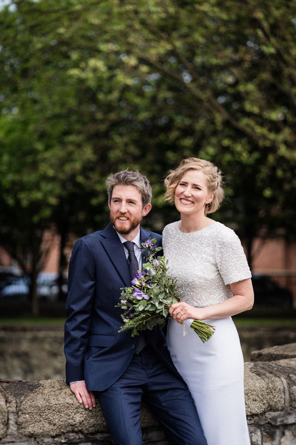 A couple poses against a stone wall with a purple floral bouquet on their wedding day.