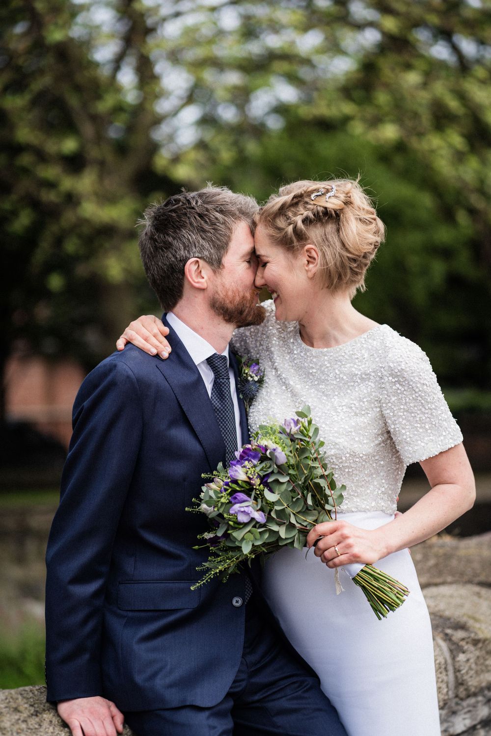 An intimate moment between newlyweds with a purple bouquet against a garden backdrop.