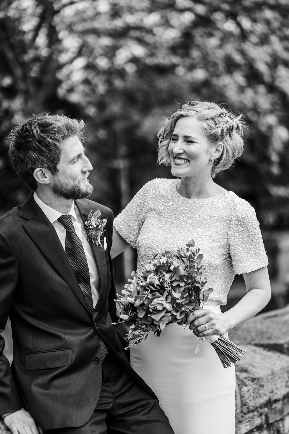 A bride in a beaded short-sleeve dress holds a colorful bouquet while sharing a joyful moment on their wedding day.