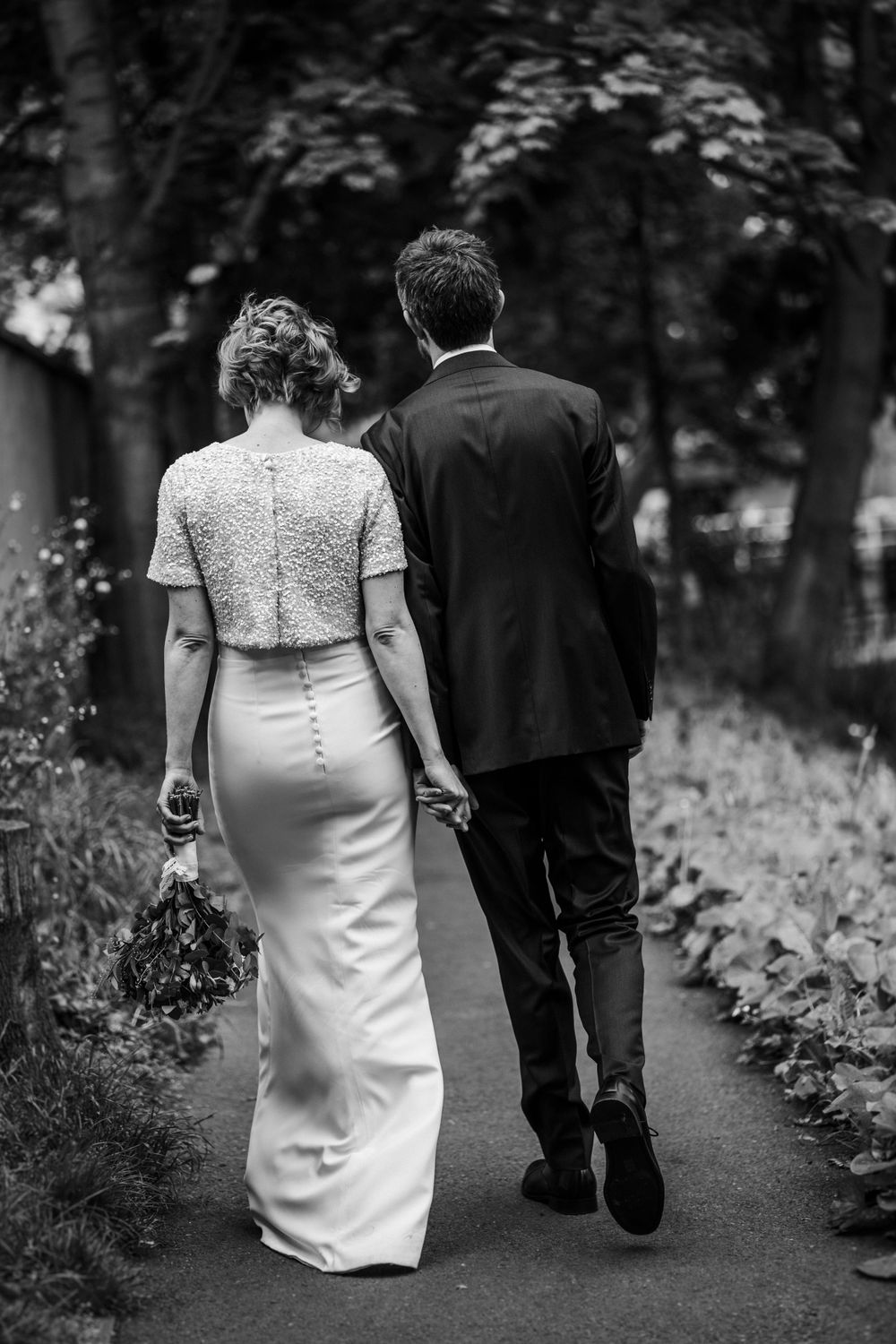 A romantic black and white photo of newlyweds walking together on a garden path.