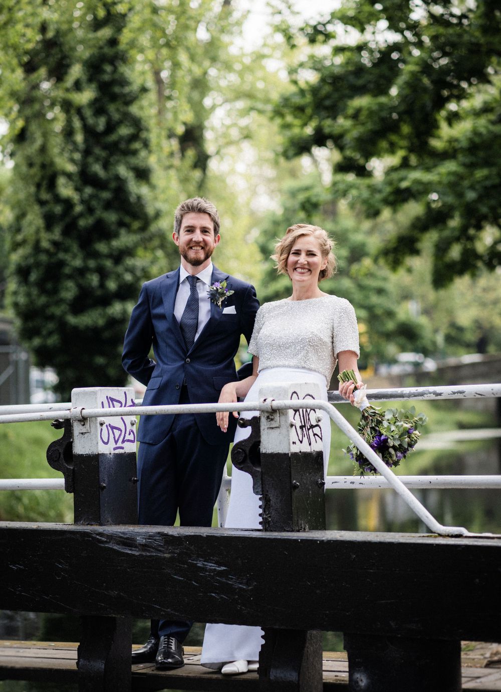 A wedding couple pose together on a wooden bridge in a scenic park setting.