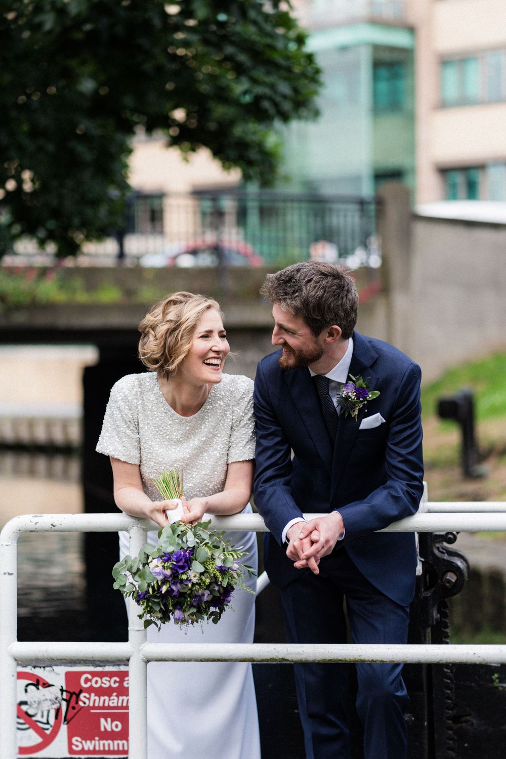A joyful wedding couple share a moment on a bridge with purple flowers.