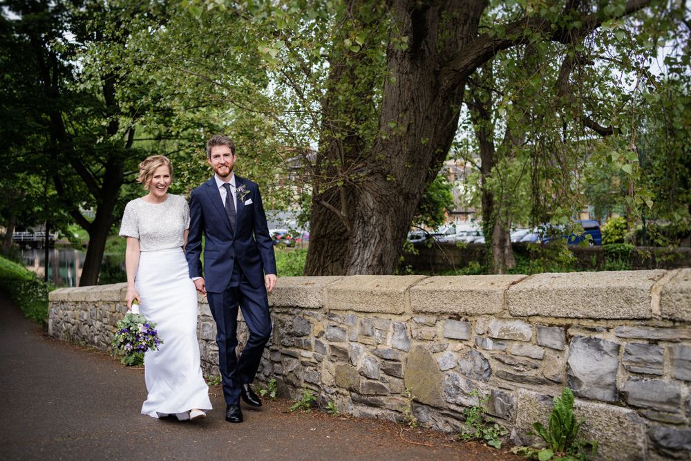 A newlywed couple walk along a stone wall path during their wedding photoshoot.