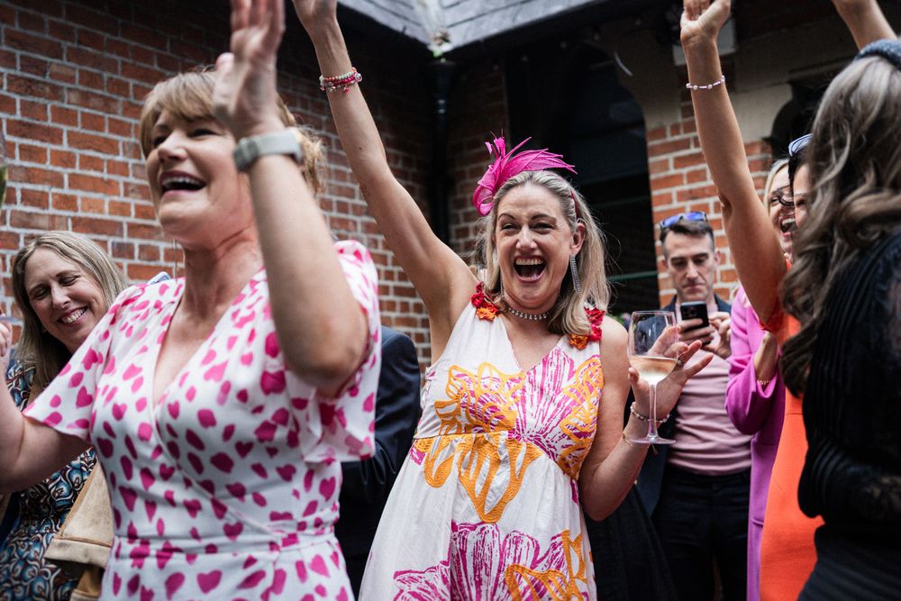 A celebration breaks out at a wedding reception with guests dancing and cheering in colorful dresses.