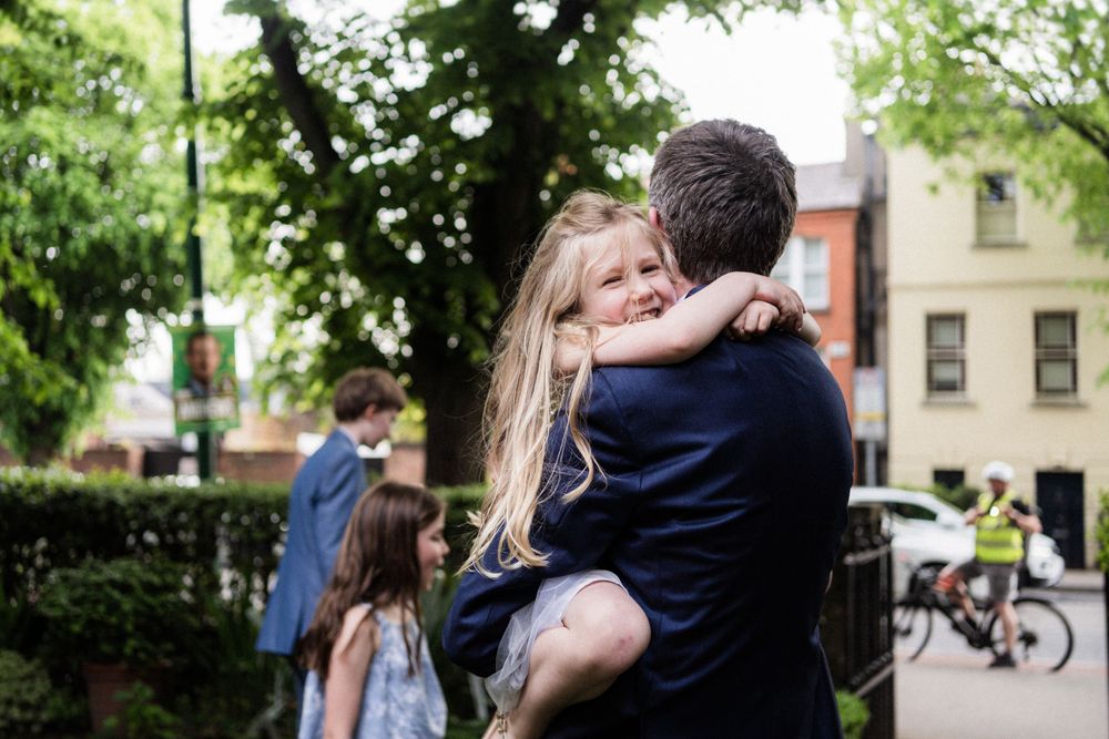 A tender moment at a wedding celebration captured in a garden setting with brick buildings in background.