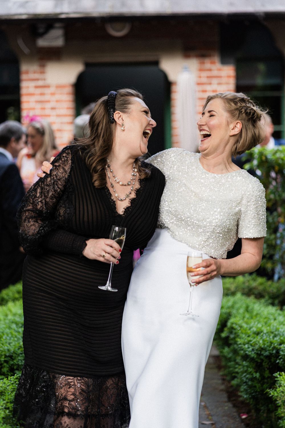 Two guests share a hearty laugh during the wedding celebration beside a brick building.